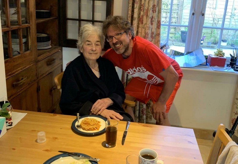 julie-sits-at-dining-table-and-philip-stands-with-arm-around-her